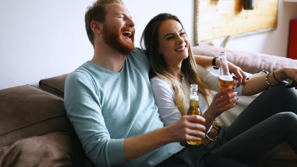A happy couple sits on a couch, laughing and holding bottles of beer.