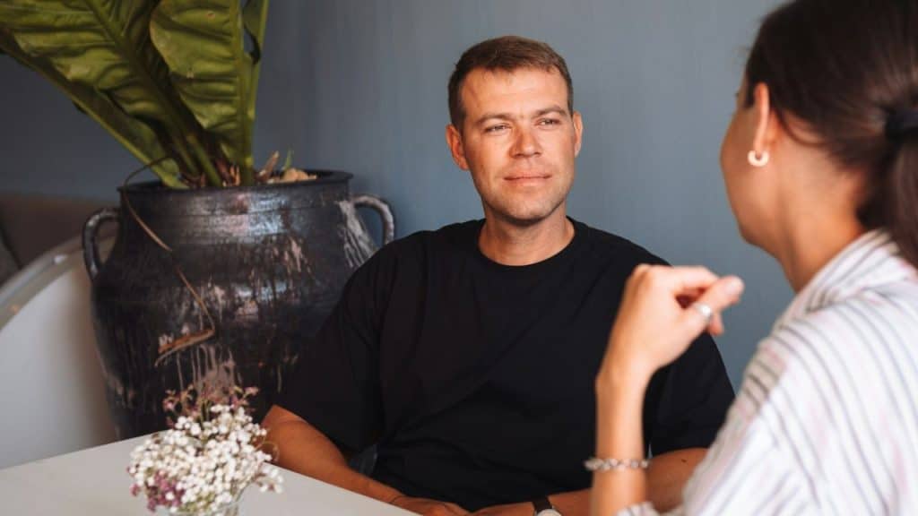 A man in a black shirt looks at a woman across a table, with a plant in a large pot nearby.