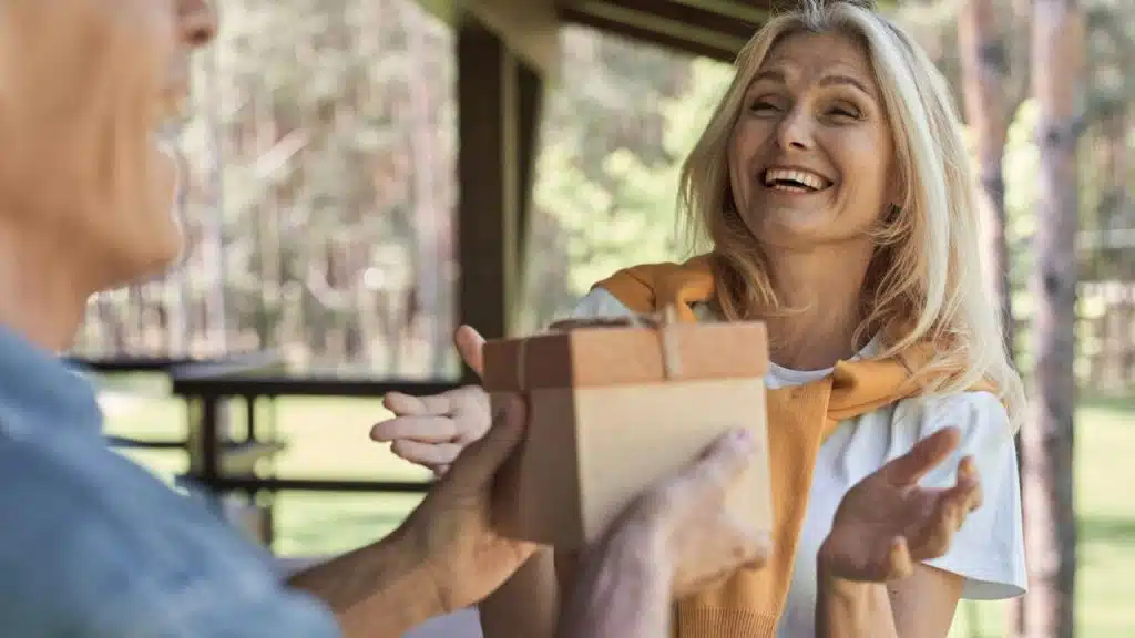 A man hands a small wrapped gift to a smiling, laughing woman outdoors.