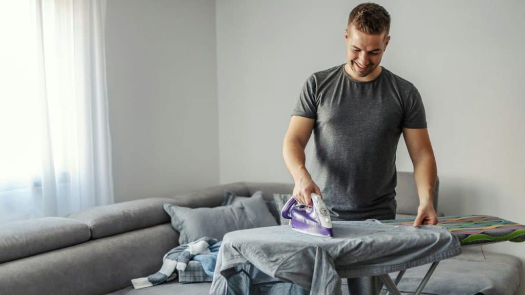 A smiling man in a grey t-shirt irons clothes on an ironing board in a living room.