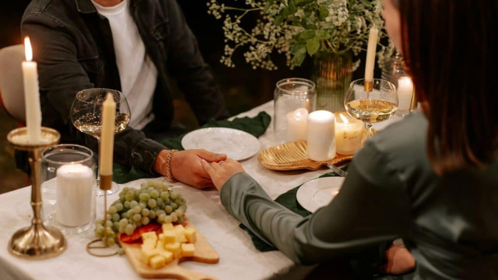 A couple holds hands across a candlelit dinner table laden with food and wine.