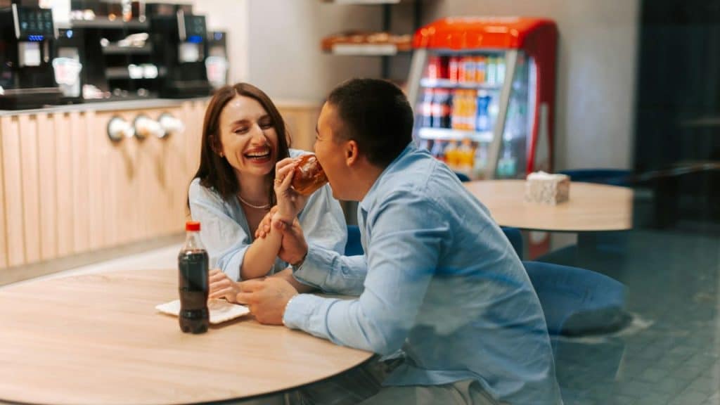 A man bites into food held by a laughing woman at a cafe table.