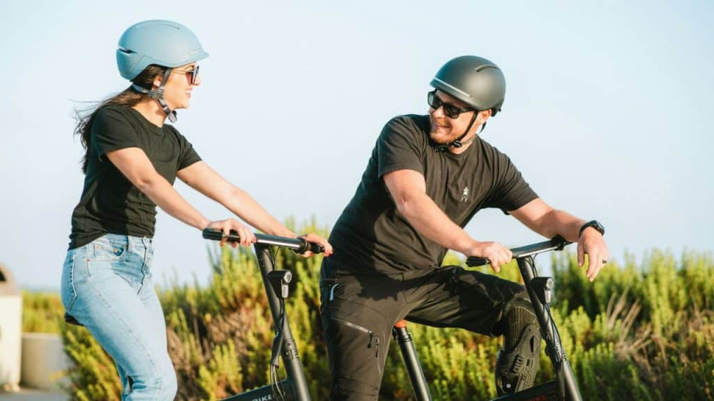 A smiling couple, wearing helmets and sunglasses, rides bicycles outdoors.