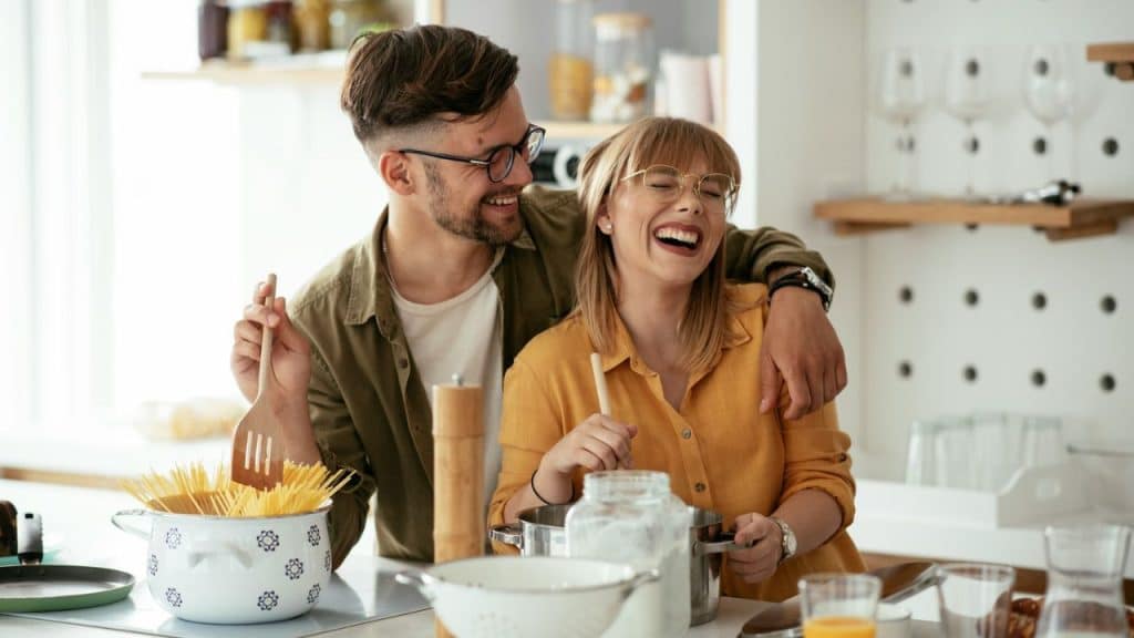 A laughing couple, wearing glasses, cooks together in a bright kitchen.