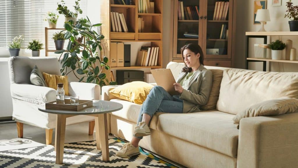 A smiling woman sits on a sofa, looking at a clipboard, in a sunny living room.