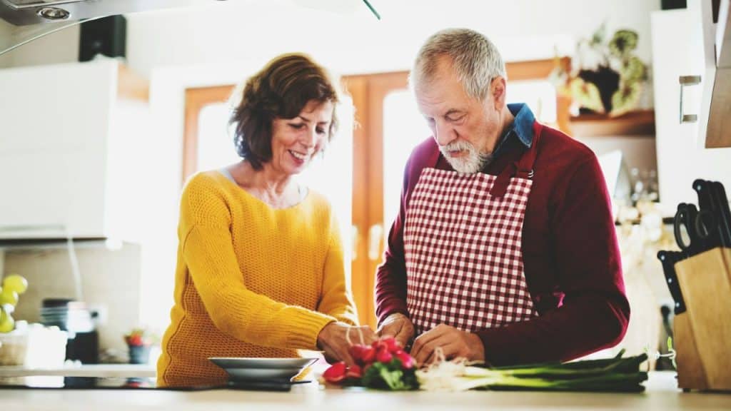 A smiling older couple prepares vegetables in a bright kitchen.