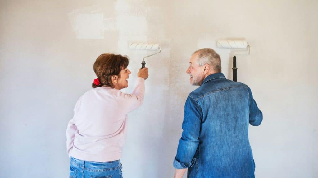 A senior couple paints a wall white, looking at each other and smiling.