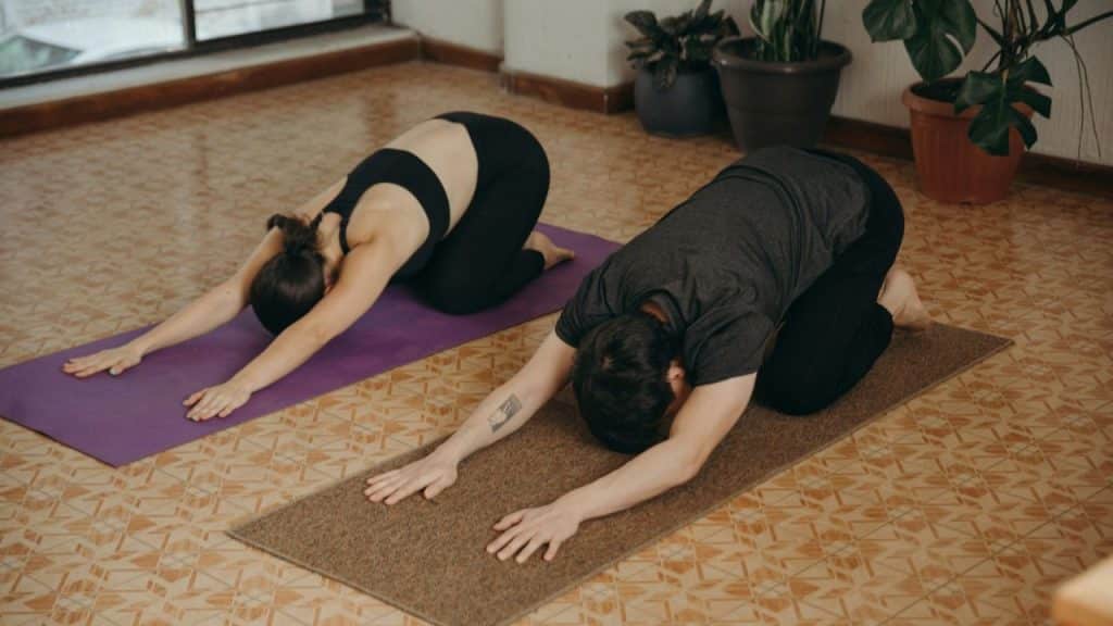 A man and woman perform the child's pose on yoga mats in a room.