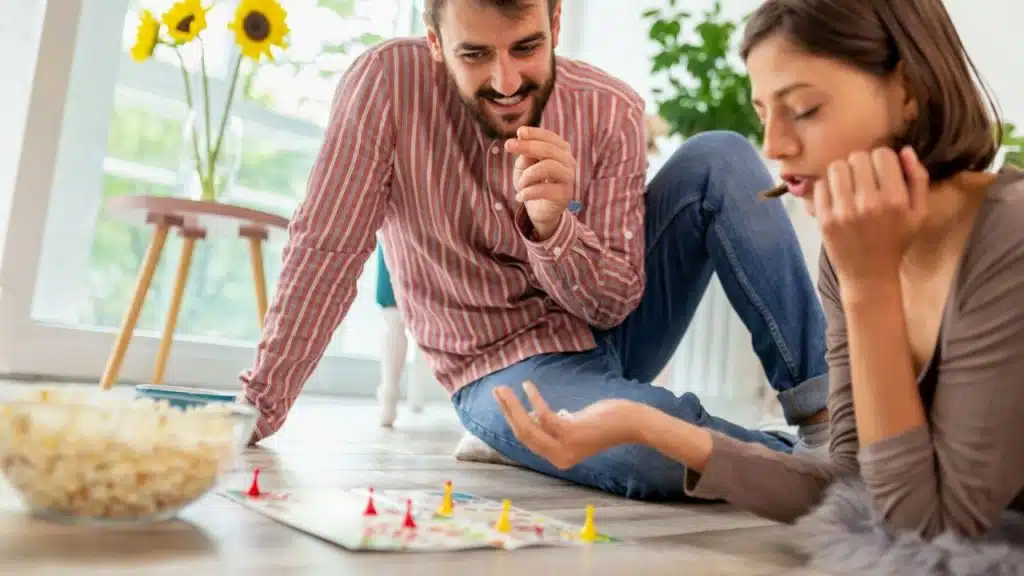 A couple sits on the floor, playing a board game with popcorn nearby.