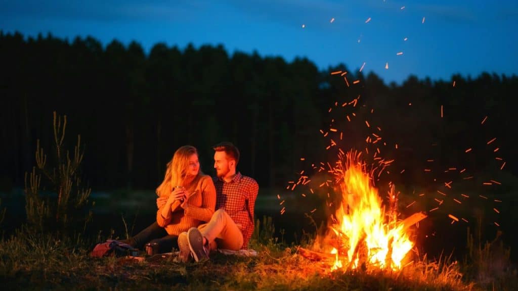 A smiling couple sits by a crackling campfire at dusk, looking at each other.