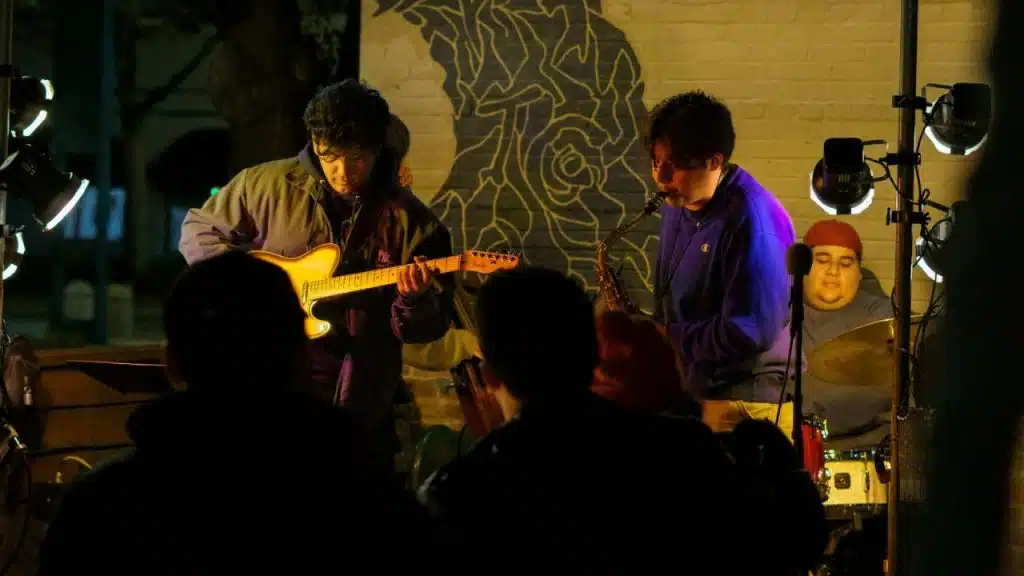 A band performs on an outdoor stage at night, with a guitarist and saxophonist in front.