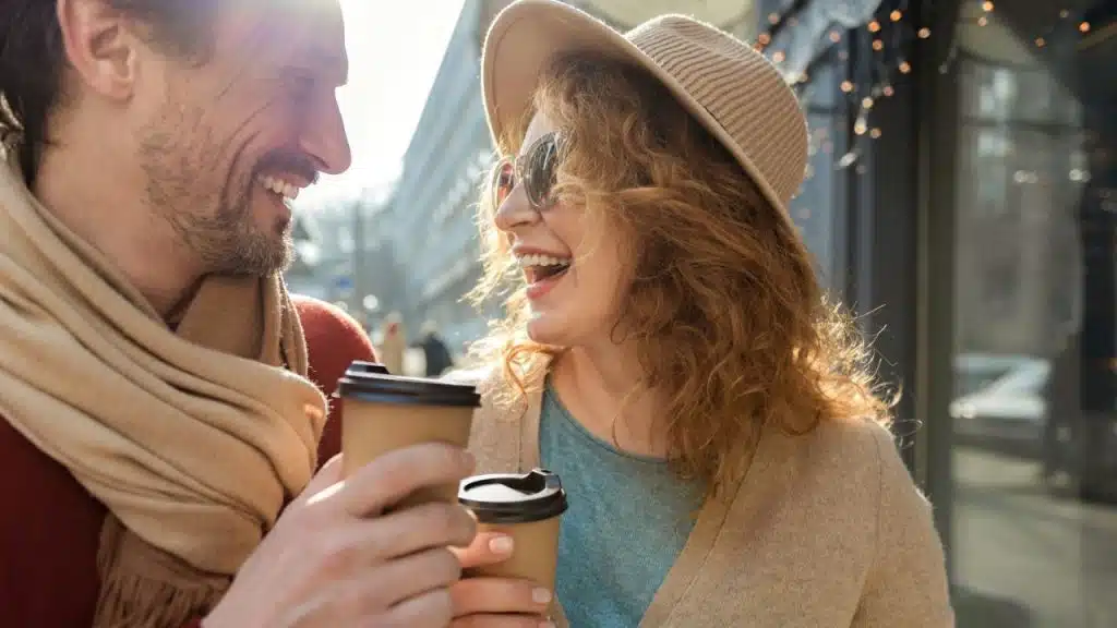 A laughing couple holds coffee cups and smiles at each other during a sunny outdoor walk.