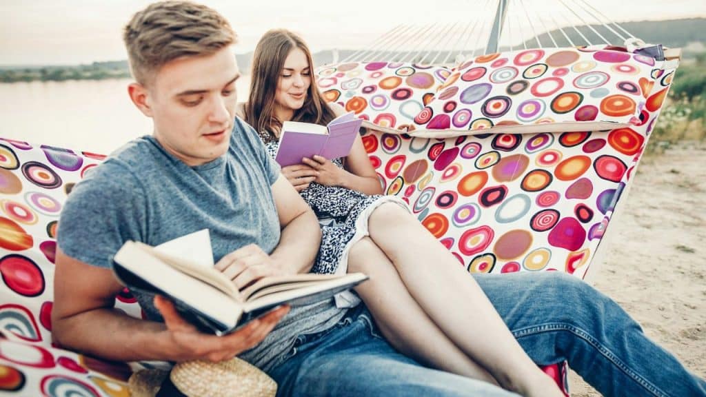 A man and woman relax in a patterned hammock, both reading books.