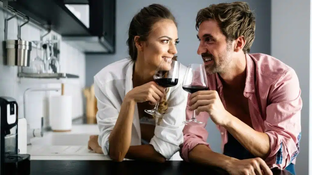 A happy couple in a kitchen smiles at each other, holding glasses of red wine.