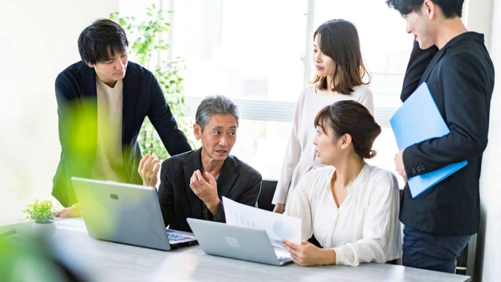 A diverse group of five professionals gathers around a table with laptops and papers, engaged in a discussion.