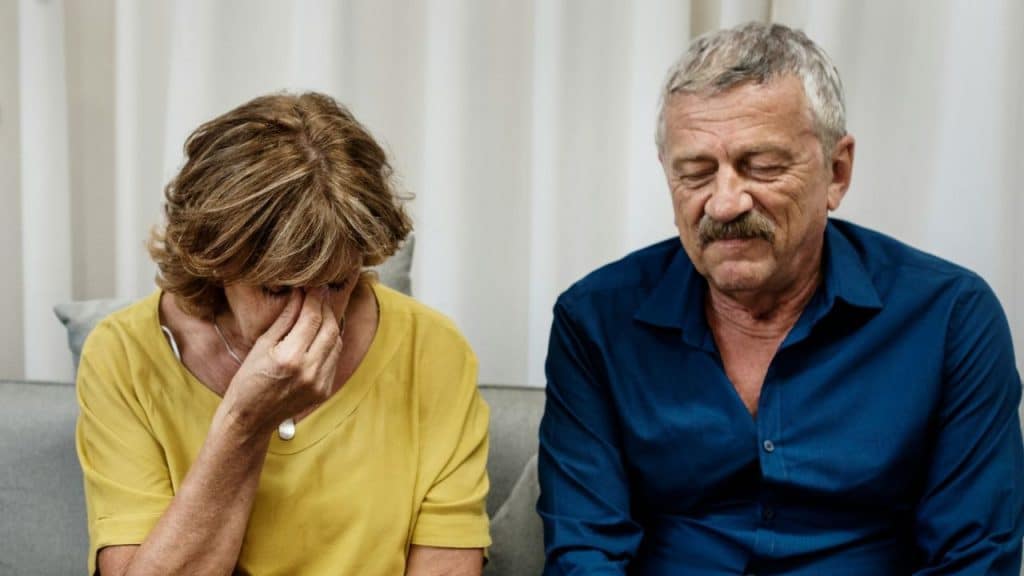 A distressed woman covers her face with her hand, sitting next to a man with his eyes closed.