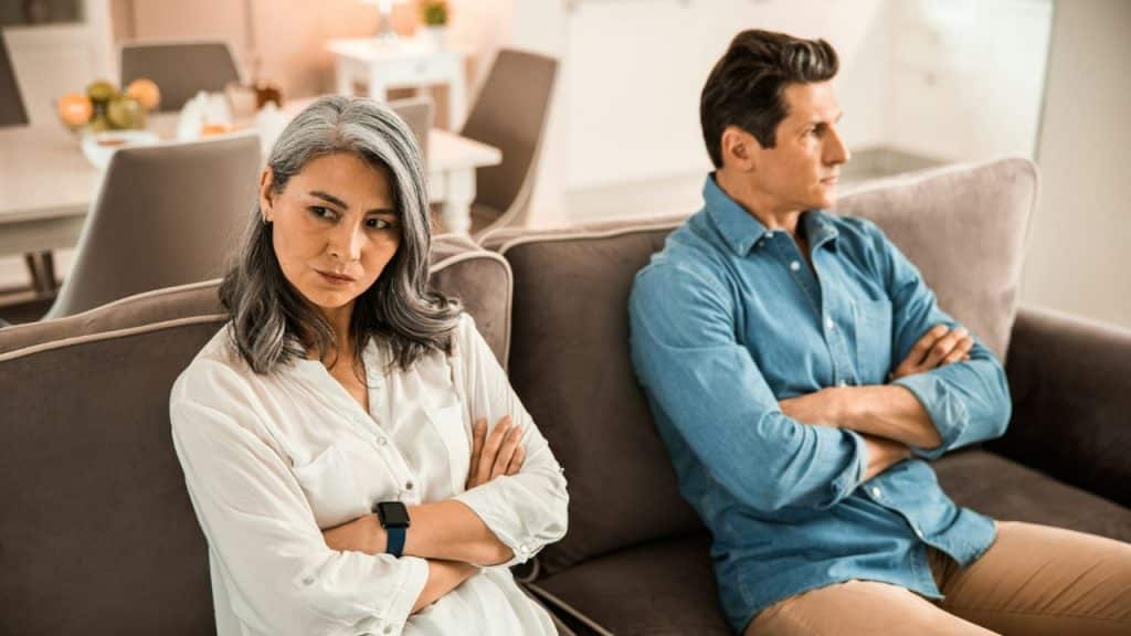 A displeased woman with grey hair and a man sit on a couch with their arms crossed, looking away from each other.