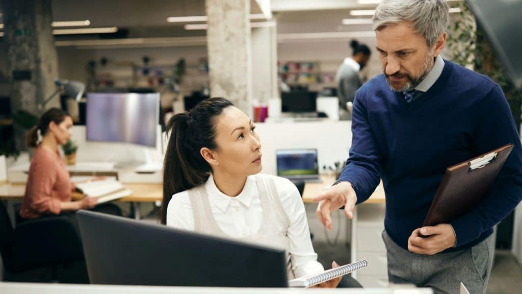 A mature man with grey hair and a beard, holding a clipboard, gestures while talking to a young Asian woman at her desk in an open office.