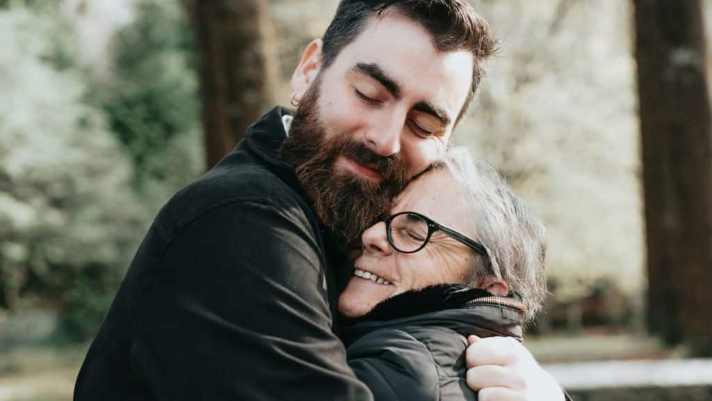 A smiling man with a beard hugs an older woman wearing glasses, both with their eyes closed.