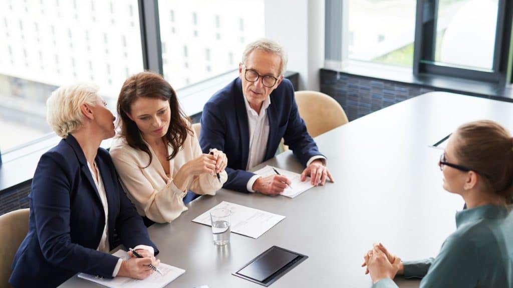 A man and two women sit at a conference table, looking at a person with their back to the camera.