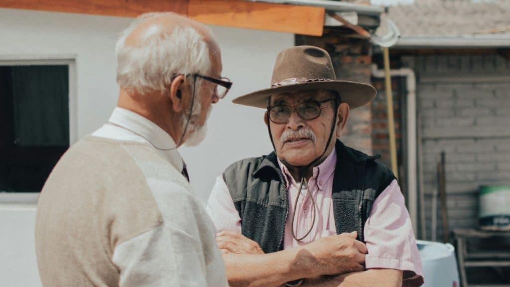 Two older men, one in a hat and glasses, look at each other and talk outdoors.