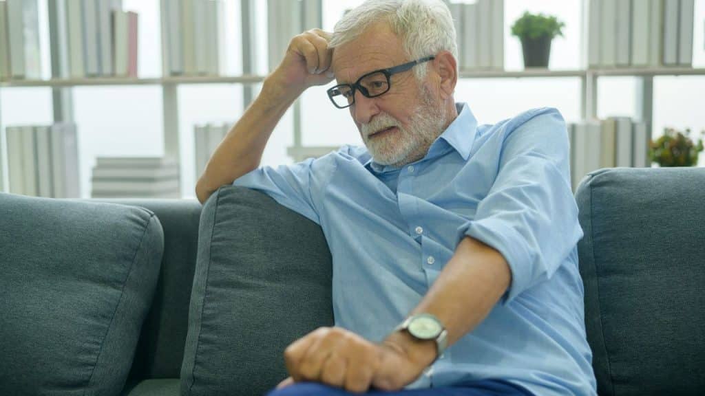 A distressed older man with glasses and a beard sits on a couch, his hand on his forehead.