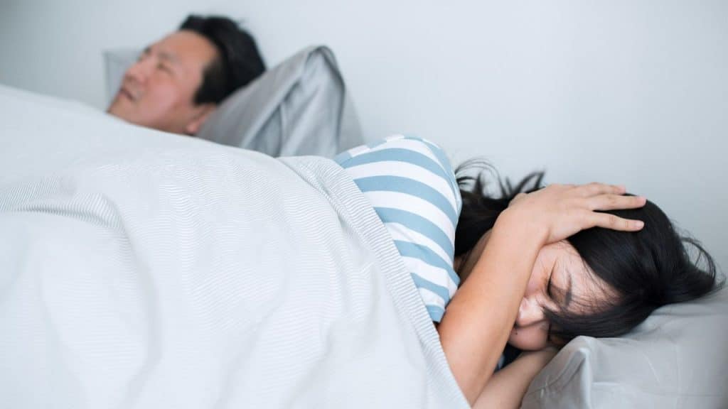 A man lies asleep under covers while a woman beside him covers her face with her hand.