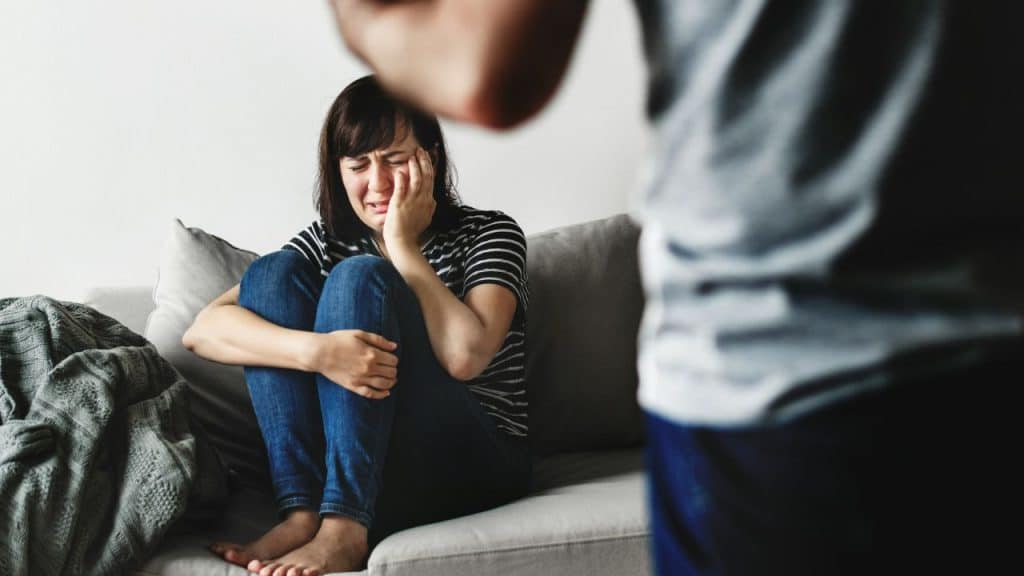 A crying woman sits on a couch, holding her face, while a blurry man stands nearby.