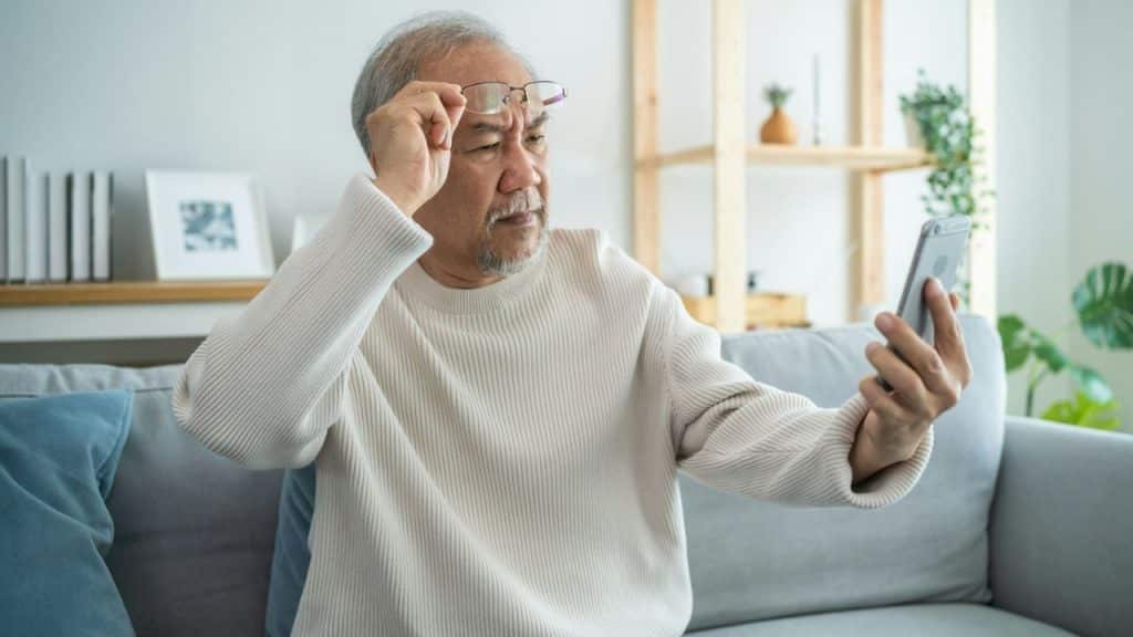 An older Asian man with a beard and glasses holds his phone away from his face, looking at it with a squint, as if having trouble seeing.