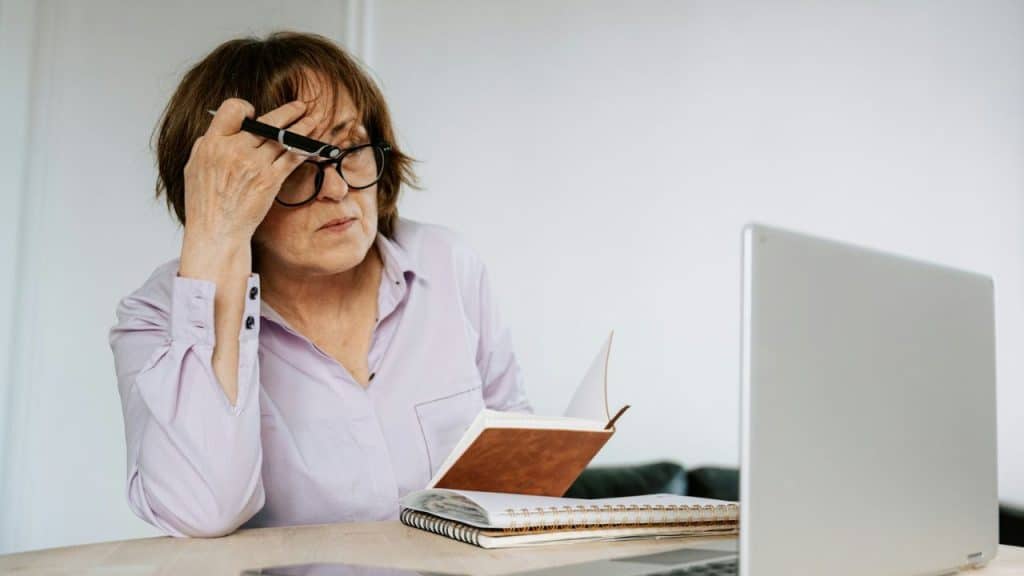 A distressed older woman wearing glasses holds a pen to her forehead while looking at a notebook with a laptop nearby.