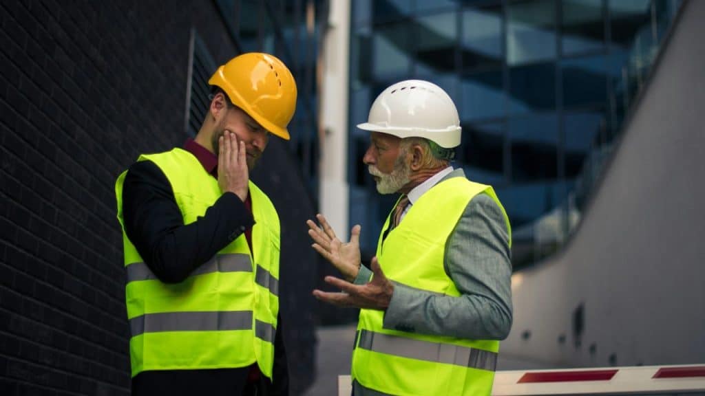 Two men in hard hats and safety vests are talking outdoors, with one man gesturing and the other holding his hand to his face.