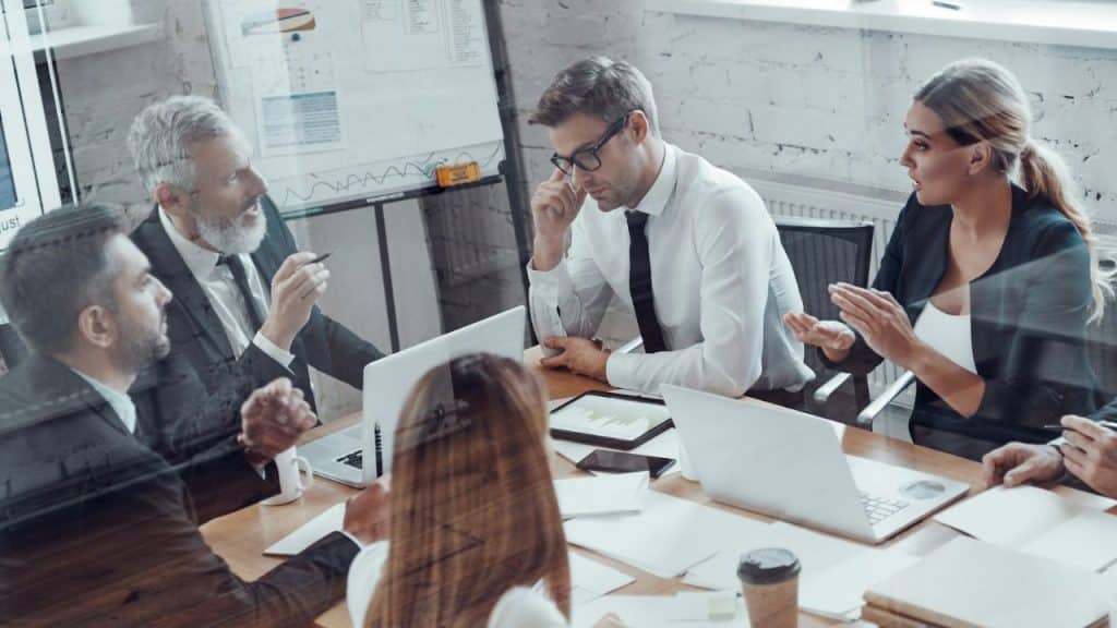 A diverse group of professionals is engaged in a discussion around a conference table with laptops and papers.