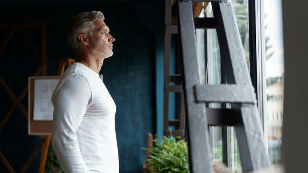 A grey-haired man in a white long-sleeved shirt stands, looking out a window.