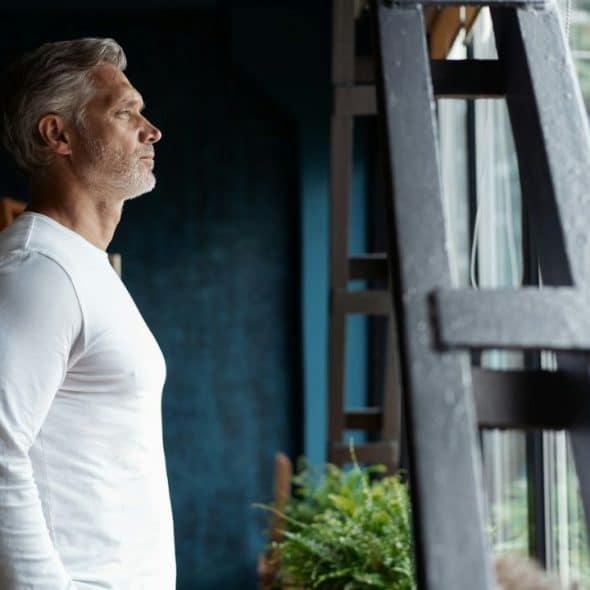 A grey-haired man in a white long-sleeved shirt stands, looking out a window.
