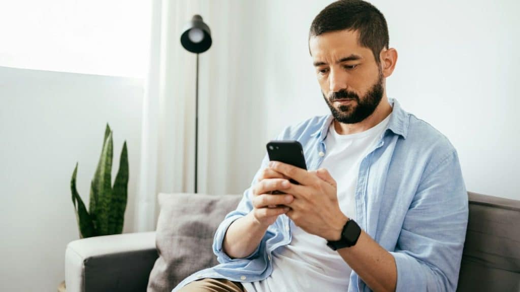 A bearded man sits on a couch, intently looking at his phone.