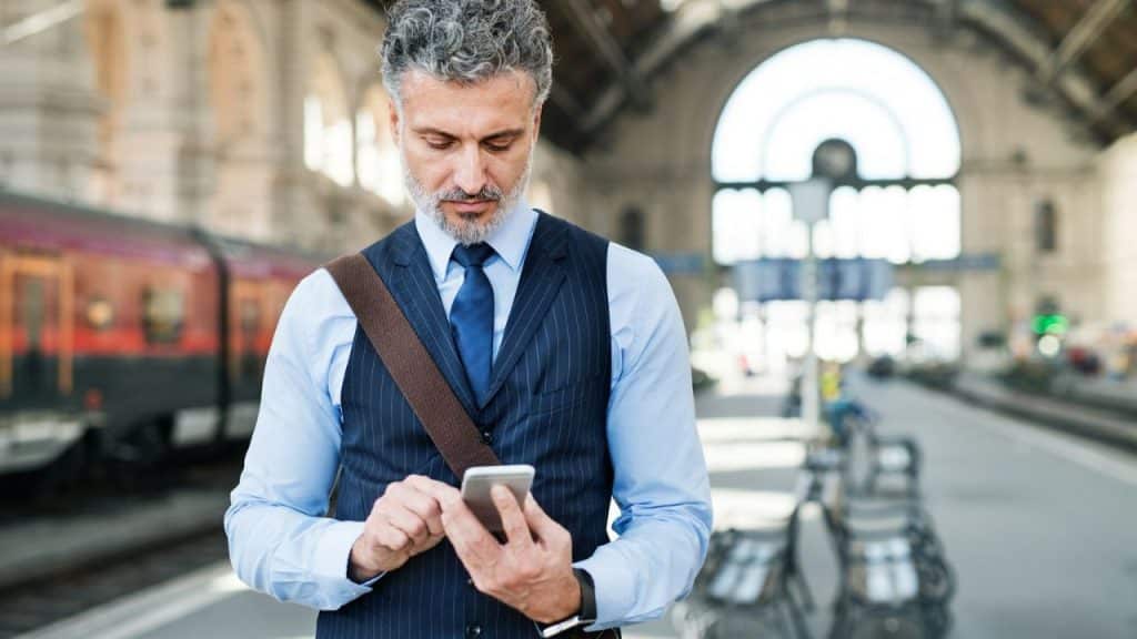 A businessman with grey hair and a beard looks at his phone while standing on a train platform.