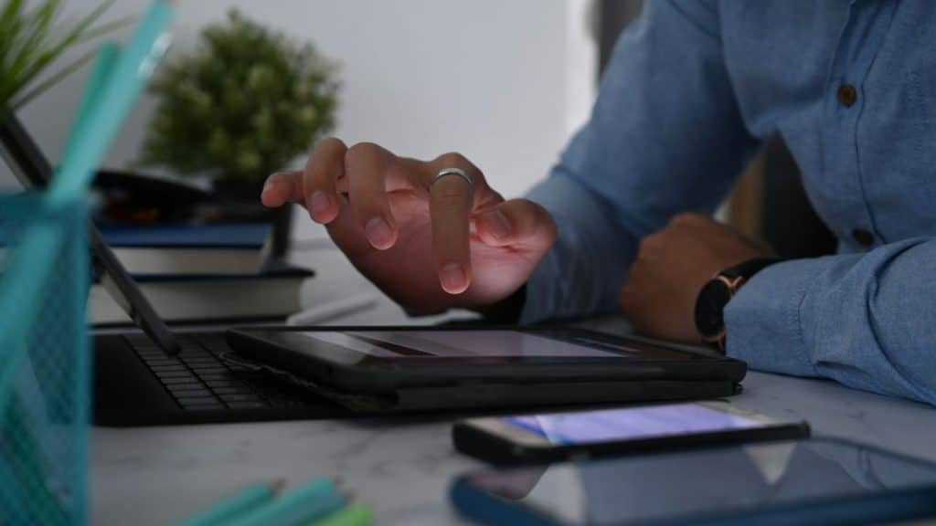 A person's hand with a ring on the index finger touches a tablet screen, with a keyboard, phone, and books nearby.