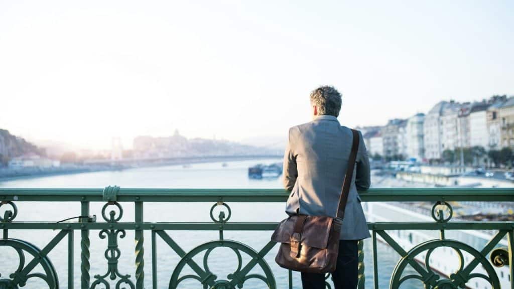 A man with a satchel over his shoulder stands on a bridge, looking out at a city and river.