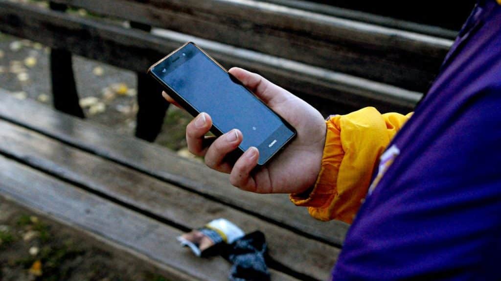 A person wearing a purple and yellow jacket holds a smartphone, with a wooden bench in the background.