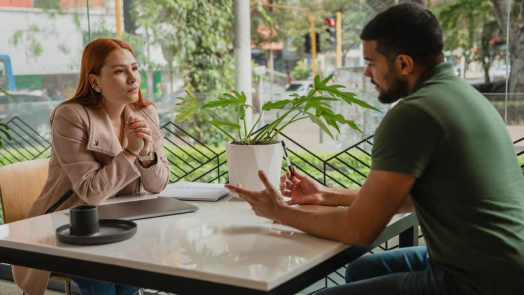 A woman and man talk across a table with a plant and laptop in a cafe.