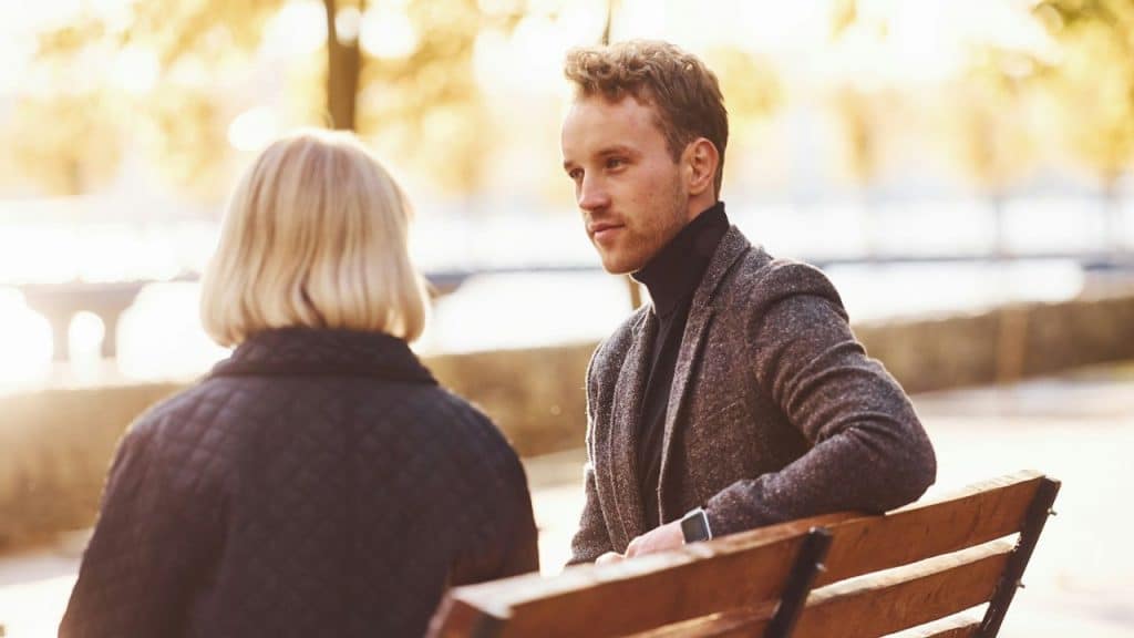 A man in a tweed jacket looks at a woman with blonde hair, sitting on a park bench.