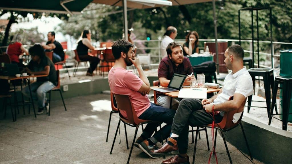 Three men sit at an outdoor table, talking and looking at a laptop, with other patrons in the background.