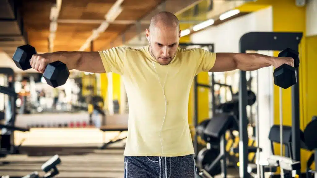 A bald man in a yellow t-shirt and earbuds is lifting dumbbells out to his sides in a gym.