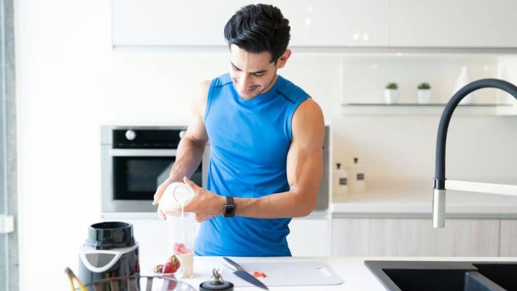 A smiling man in a blue tank top pours liquid from a container into a cup in a modern kitchen.