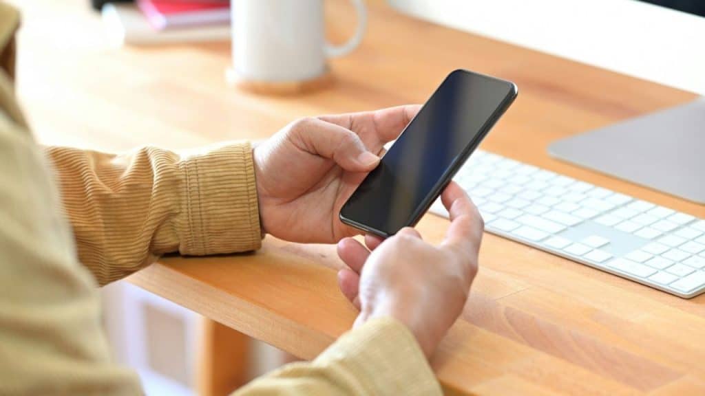 A person holds a black smartphone with both hands, near a computer keyboard and mug on a wooden desk.