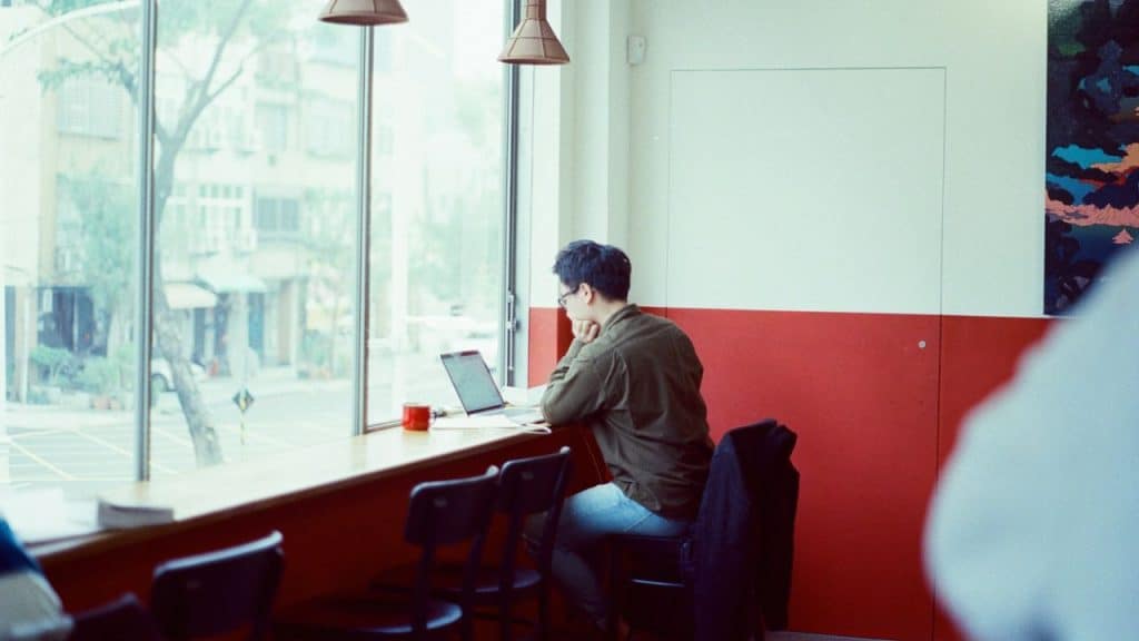 A man sits at a window counter with a laptop and a red mug, looking out at a street.