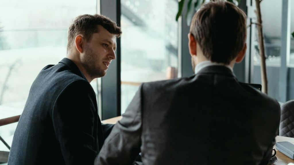 Two men in suits are seated at a table, one looking at the other while the second man has his back to the camera.