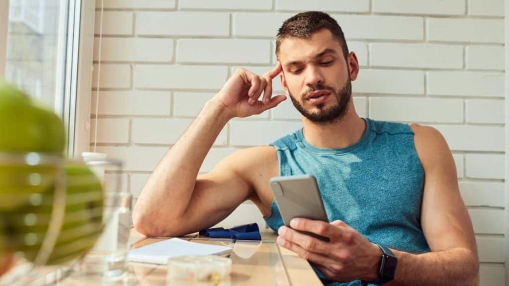 A man in a blue tank top looks at his phone with a thoughtful expression, his hand on his temple, at a table near a window.