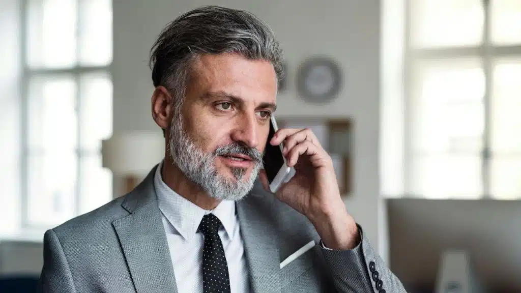 A grey-haired man in a suit and tie talks on a smartphone, looking thoughtful.