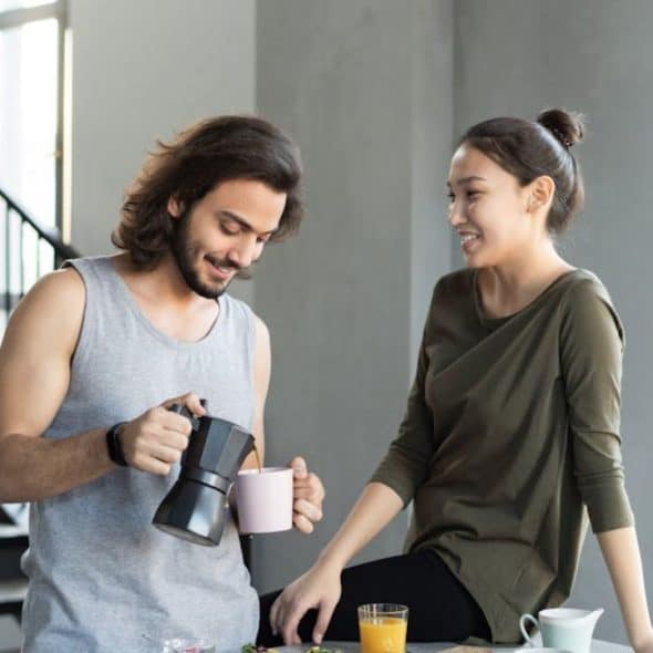 A man smiling while handing his partner a coffee in the kitchen during a relaxed morning.